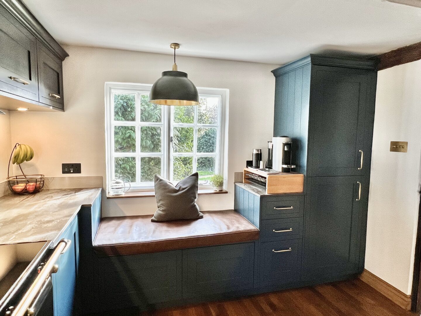Painted Shaker style blue kitchen with Brass handles, cornice and induction Rangecooker with brass knobs, walnut effect wall units and Dekton worktops and a window seat