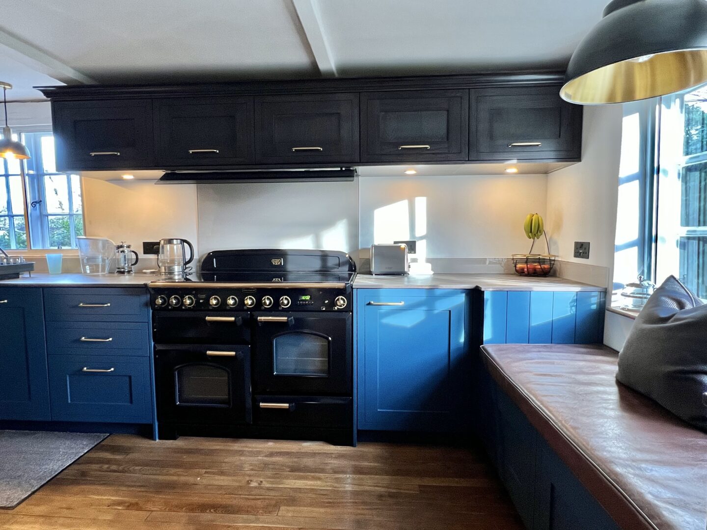 Bespoke Shaker Kitchen in Sandhurst. Shaker style blue kitchen with Brass handles, cornice and induction Rangecooker with brass knobs, walnut effect wall units and Dekton worktops and a window seat